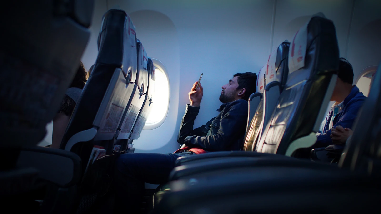 Passenger in a window seat on an airplane looking at a smartphone in soft blue cabin light