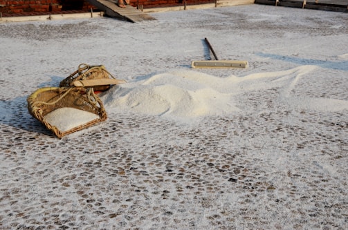 Baskets filled with rice are placed on a textured surface, possibly a drying area. A wooden tool lies nearby, suggesting ongoing work or a recent task. Sunlight casts soft shadows, adding depth to the scene.