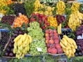 A vibrant assortment of fruits displayed in a market setting. There are bunches of bananas, grapes, plums, peaches, cherries, and apricots neatly arranged in sections. The fruits are fresh and colorful, with greenery interspersed among them, adding to the visual appeal.