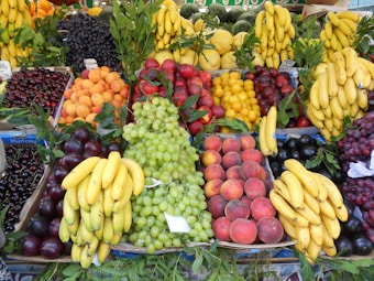 A vibrant assortment of fruits displayed in a market setting. There are bunches of bananas, grapes, plums, peaches, cherries, and apricots neatly arranged in sections. The fruits are fresh and colorful, with greenery interspersed among them, adding to the visual appeal.