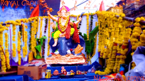 A vibrantly decorated statue of the Hindu deity Ganesha sits on an intricately adorned platform, surrounded by yellow marigold garlands. An elephant head forms part of the statue's base, and the setup includes decorative leaves and offerings. Boxes and other items are placed nearby.