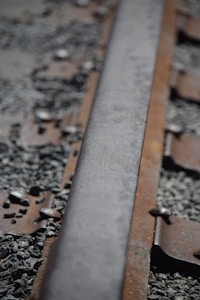 A close-up of a section of weathered railway track with rusty metal and scattered gravel surrounding it. The perspective focuses on a single rail, highlighting its texture and the wear over time.