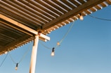 A wooden pergola with evenly spaced slats casts shadows against a clear blue sky. Light bulbs hang from green cords attached to the structure. A bird perches on one of the cords, adding a natural element to the scene.