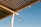 A wooden pergola with evenly spaced slats casts shadows against a clear blue sky. Light bulbs hang from green cords attached to the structure. A bird perches on one of the cords, adding a natural element to the scene.