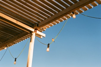 A wooden pergola with evenly spaced slats casts shadows against a clear blue sky. Light bulbs hang from green cords attached to the structure. A bird perches on one of the cords, adding a natural element to the scene.
