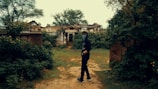 A person stands on a dirt path surrounded by dense greenery and overgrown vegetation. In the background, there are ruins of an old building, partially obscured by the foliage. The individual wears a dark outfit with a face covering, giving an air of mystery.