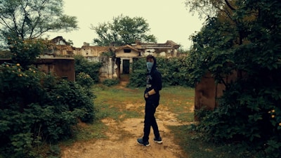 A person stands on a dirt path surrounded by dense greenery and overgrown vegetation. In the background, there are ruins of an old building, partially obscured by the foliage. The individual wears a dark outfit with a face covering, giving an air of mystery.