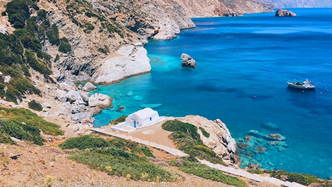 A small white building is situated on a rocky, green-tinged hillside overlooking crystal-clear turquoise waters. A boat is anchored nearby in the serene blue sea. The rocky coastlines and sparse vegetation depict a dry, yet beautiful Mediterranean landscape.