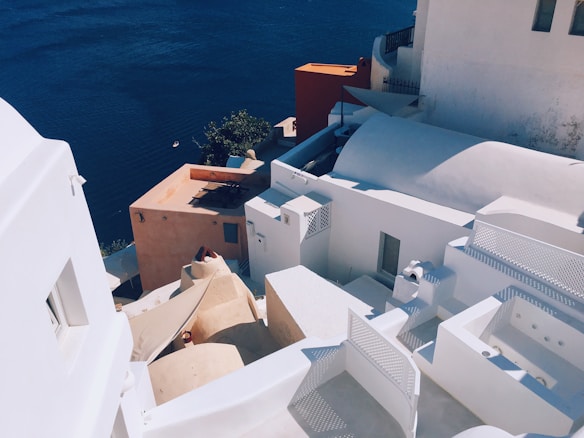 A picturesque view of whitewashed buildings with domed roofs, terraced against a backdrop of deep blue sea. The structures exhibit a traditional architectural style, with smooth curves and contrasting earthy tones. Bright sunlight casts shadows, highlighting the minimalistic design and texture of the surfaces.