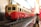 A model train painted in red and cream colors is positioned on a set of miniature tracks. The train is detailed, with small windows and intricate design elements, set against a blurred background creating a sense of depth.