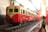 A model train painted in red and cream colors is positioned on a set of miniature tracks. The train is detailed, with small windows and intricate design elements, set against a blurred background creating a sense of depth.