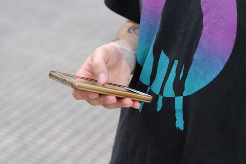 A person holding a smartphone in their hand, wearing a black shirt with a graphic design featuring a silhouette of an animal against a gradient circle in blue and purple tones.