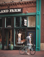 A man stands on a brick sidewalk with a bicycle, looking at his phone in front of a clothing store called Island Farm. The storefront features large windows displaying mannequins with casual clothing. The building facade is brick with green and teal trim accents, and potted plants flank the entrance.