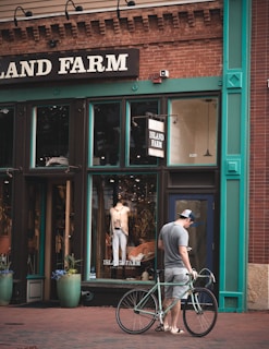 A man stands on a brick sidewalk with a bicycle, looking at his phone in front of a clothing store called Island Farm. The storefront features large windows displaying mannequins with casual clothing. The building facade is brick with green and teal trim accents, and potted plants flank the entrance.