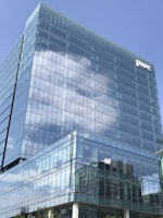 A modern glass office building reflecting the blue sky and clouds in its windows. The structure showcases a sleek and symmetrical design. A company logo is visible on the top corner of the building.