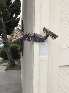 A set of handmade directional signs made from cardboard are attached to the corner of a building. The signs contain messages indicating an 'Alley Sale' happening on Saturday and that it will be 'Awesome.' The building is off-white, and there is a tree and some bushes nearby.