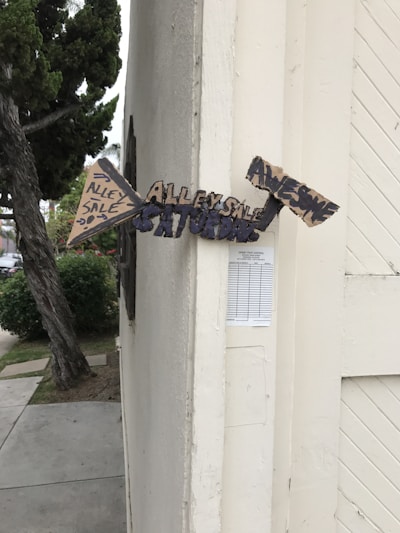 A set of handmade directional signs made from cardboard are attached to the corner of a building. The signs contain messages indicating an 'Alley Sale' happening on Saturday and that it will be 'Awesome.' The building is off-white, and there is a tree and some bushes nearby.