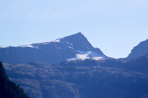 A rugged mountain landscape with a prominent peak partially covered by patches of snow. The jagged outlines of the mountains are contrasted against a light blue sky. Shadows and textures on the rocky surfaces highlight the uneven terrain, while sparse vegetation appears on the lower slopes.