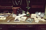 An antique wooden desk is adorned with vintage items including old photographs, a pair of black binoculars, a brown leather case, a marble with a wooden base, a decorative knife, and a small vase with flowers. The background consists of shelves filled with envelopes and papers.