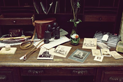 An antique wooden desk is adorned with vintage items including old photographs, a pair of black binoculars, a brown leather case, a marble with a wooden base, a decorative knife, and a small vase with flowers. The background consists of shelves filled with envelopes and papers.