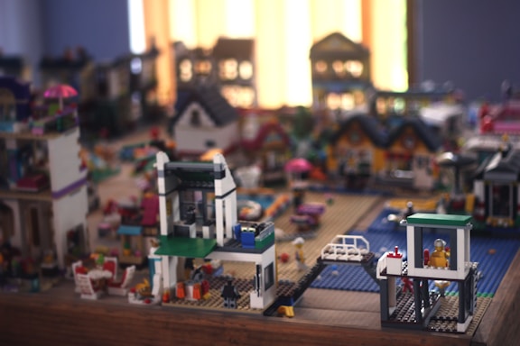 A detailed miniature cityscape built from colored toy bricks, featuring various buildings and structures. Bright window light illuminates the scene, creating a cozy atmosphere. There are houses with sloped roofs, a small balcony with chairs, and a blue base implying water. Toy figures are visible, enhancing the lively setting.