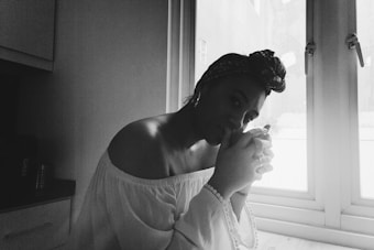 A person sits by a window, holding a cup with both hands. The lighting is soft, casting a gentle shadow on the person&rsquo;s face. They are wearing an off-the-shoulder top and a headscarf. The scene has a relaxed and contemplative atmosphere.