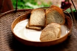 A wooden basket contains a few slices of toasted bread on a white ceramic plate. The setting has a rustic and natural feel, with an out-of-focus background of green foliage.
