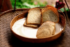 A wooden basket contains a few slices of toasted bread on a white ceramic plate. The setting has a rustic and natural feel, with an out-of-focus background of green foliage.