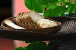 A wicker tray holds a wooden plate filled with neatly arranged slices of bread. Fresh green leaves in the background provide a natural and earthy setting.