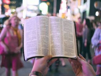 person holding bible on road with people walking on sidewalk beside buildings during nighttime