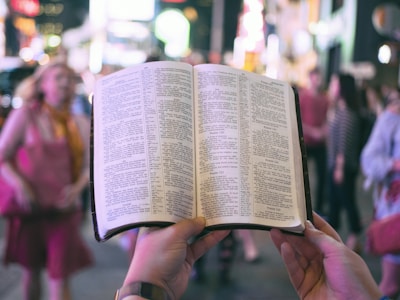 person holding bible on road with people walking on sidewalk beside buildings during nighttime