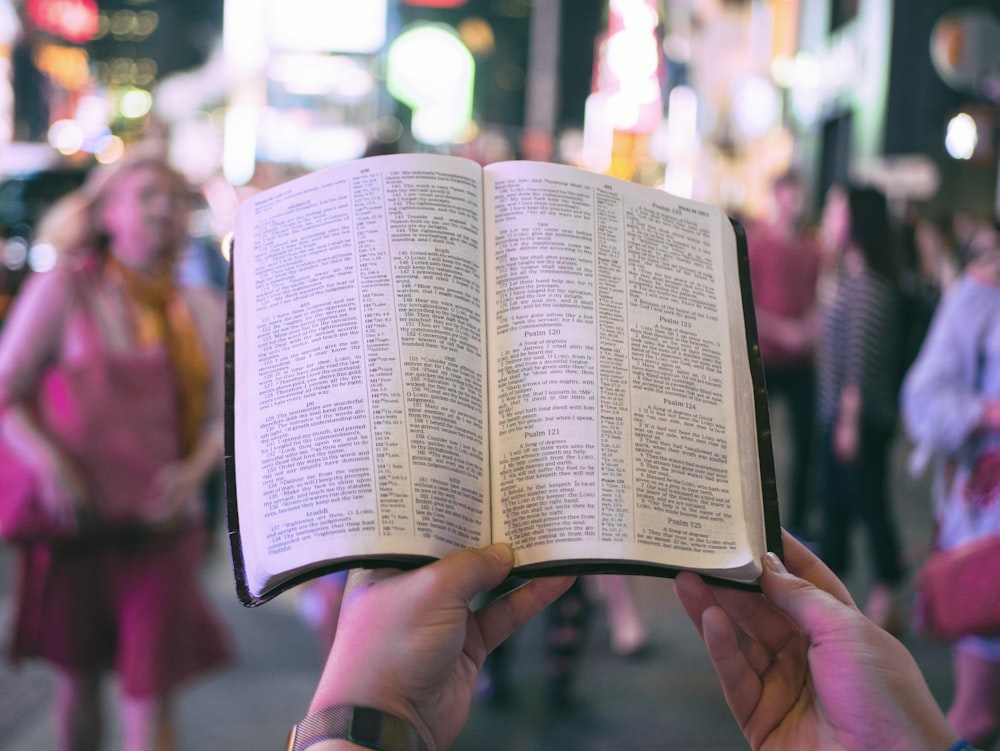 person holding bible on road with people walking on sidewalk beside buildings during nighttime