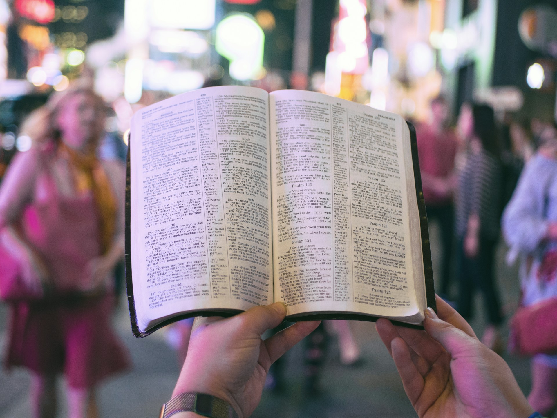 person holding bible on road with people walking on sidewalk beside buildings during nighttime