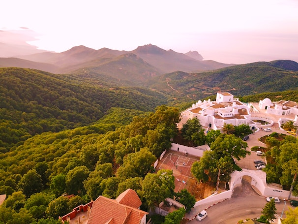 A scenic view of a lush green forested landscape with rolling hills and mountains in the background. In the foreground, a cluster of white buildings with red-tiled roofs is visible, surrounded by greenery. The winding road and parked cars lead to the entrance of the complex, emphasizing the harmony between architecture and nature.