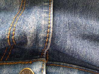 Close-up view of blue denim fabric featuring orange stitching details and a metal button.
