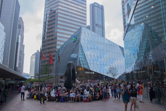 A bustling urban scene featuring crowds of people gathered around a large black abstract sculpture. Surrounding the area are modern high-rise buildings with reflective glass surfaces, some adorned with flags. The atmosphere is lively and busy as people appear to be socializing, walking, and resting.