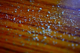 Small white granules scattered across a textured wooden surface with a warm orange-brown tone. The image is focused closely on the granules, highlighting their irregular shapes and reflective qualities. The background is subtly blurred, creating a sense of depth.
