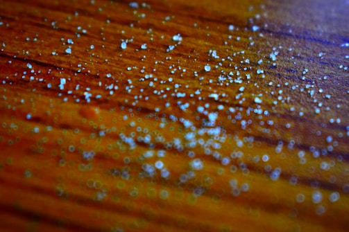 Small white granules scattered across a textured wooden surface with a warm orange-brown tone. The image is focused closely on the granules, highlighting their irregular shapes and reflective qualities. The background is subtly blurred, creating a sense of depth.