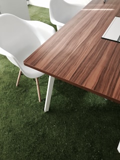 A modern wooden table with a smooth, striped pattern is situated on a surface of artificial green grass. Adjacent to the table, there are white plastic chairs with wooden legs. The setting suggests an indoor area with an emphasis on combining natural elements and minimalistic design.