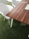 A modern wooden table with a smooth, striped pattern is situated on a surface of artificial green grass. Adjacent to the table, there are white plastic chairs with wooden legs. The setting suggests an indoor area with an emphasis on combining natural elements and minimalistic design.