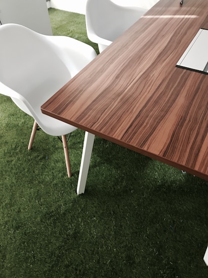 A modern wooden table with a smooth, striped pattern is situated on a surface of artificial green grass. Adjacent to the table, there are white plastic chairs with wooden legs. The setting suggests an indoor area with an emphasis on combining natural elements and minimalistic design.