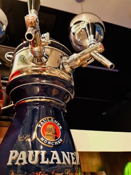 A close-up view of a beer tap with shiny chrome surfaces, featuring the logo of Paulaner München on a dark blue background. The image showcases the intricate details of the tap handles and the reflective surface of the equipment, with a background that includes reflective metallic elements.