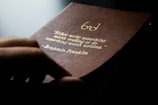 A close-up of a hand holding a brown leather-bound book with an engraved gold quote by Benjamin Franklin, emphasizing the value of writing and reading.