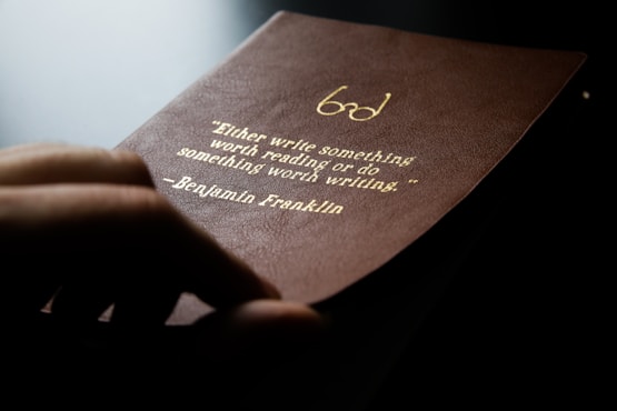 A close-up of a hand holding a brown leather-bound book with an engraved gold quote by Benjamin Franklin, emphasizing the value of writing and reading.