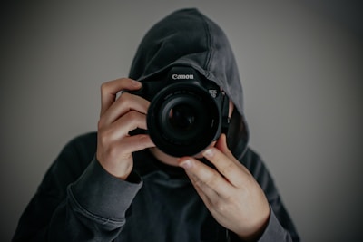 A person wearing a dark hoodie holds a Canon camera up to their face, obscuring their facial features. The image is focused on the camera lens, creating a mysterious and anonymous feel.