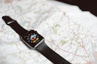 A smartwatch with a black band is resting on an unfolded map. The smartwatch screen displays a cartoon character pointing at the clock hands.