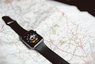 A smartwatch with a black band is resting on an unfolded map. The smartwatch screen displays a cartoon character pointing at the clock hands.