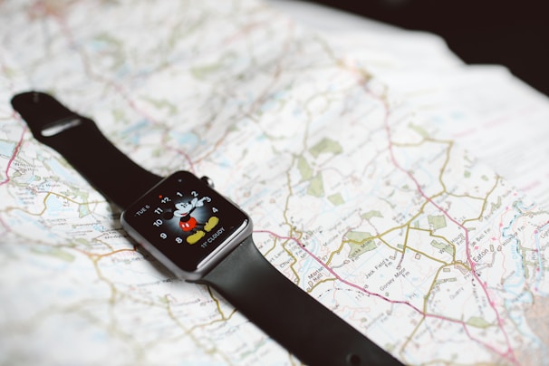 A smartwatch with a black band is resting on an unfolded map. The smartwatch screen displays a cartoon character pointing at the clock hands.