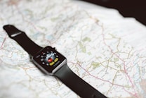 A smartwatch with a black band is resting on an unfolded map. The smartwatch screen displays a cartoon character pointing at the clock hands.