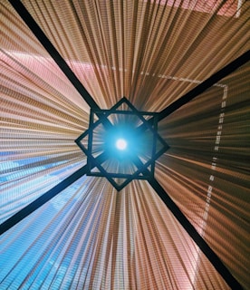 A captivating view from below of a geometric lampshade with a central light source. The shade features a symmetrical star-shaped design surrounded by pleated fabric panels that radiate outward, creating a dynamic pattern of light and shadow.